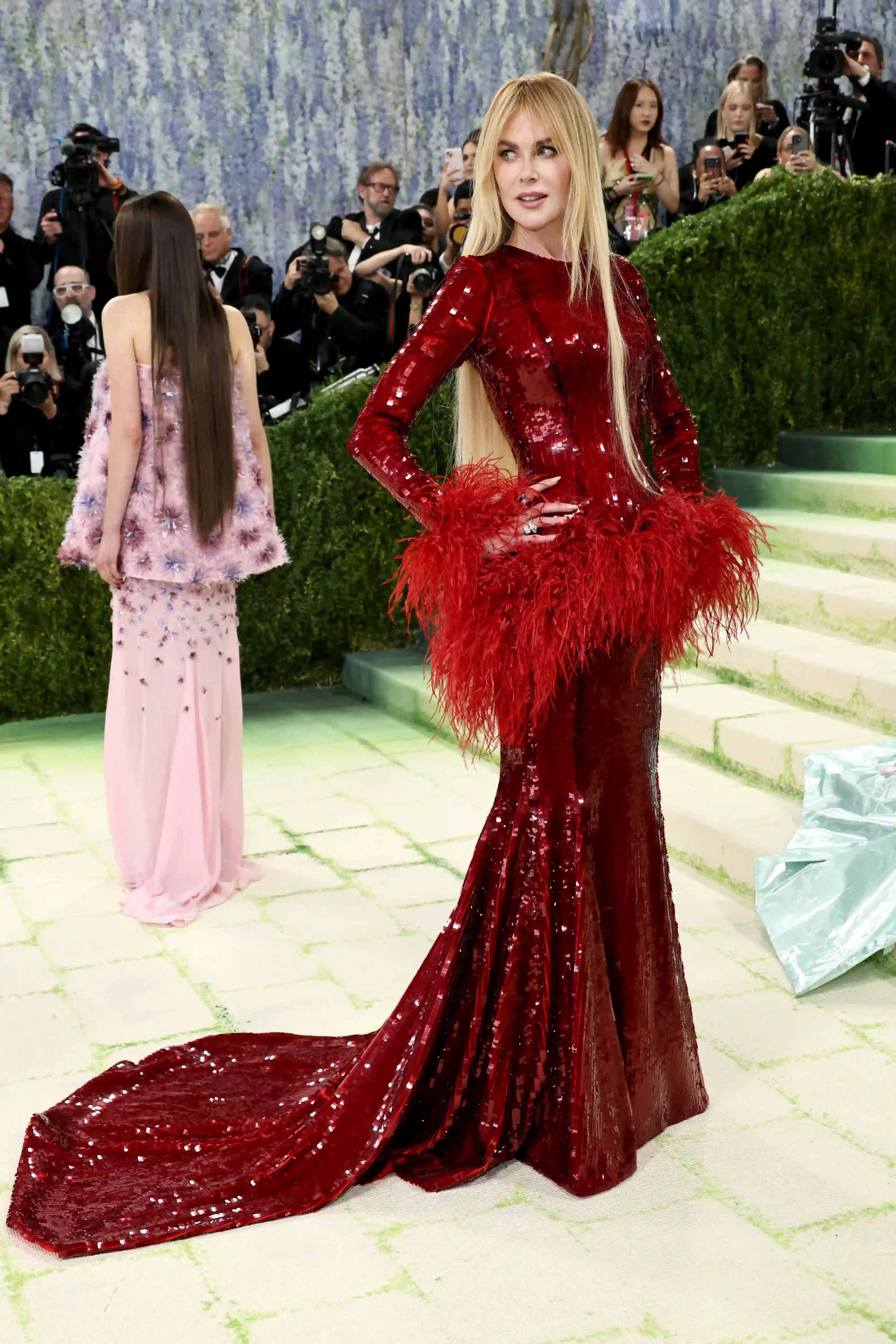 Nicole Kidman, 2026 Met Gala kırmızı halısında, baştan aşağı kırmızı pullarla işlenmiş, bel kısmında kabarık kırmızı tüylerle zenginleştirilmiş, uzun kuyruklu ve ışıltılı elbisesiyle dikkat çekici bir stil sergilerken, uzun sarı saçları ve parmağındaki yüzüklerle poz veriyor; arka planda uzun koyu saçlı başka bir katılımcı ve fotoğrafçılardan oluşan kalabalık, yeşil desenli açık renkli bir zemin üzerinde beliriyor.
