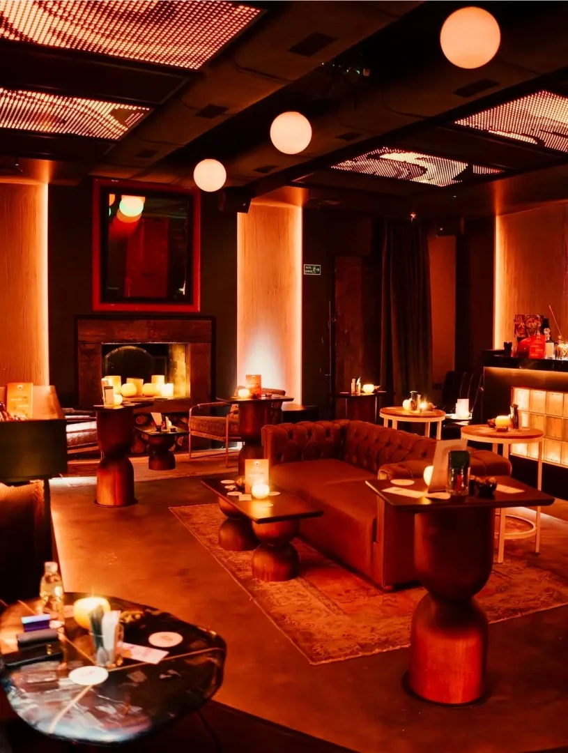 Şık, kızıl-turuncu tonlarında sıcak ışıklarla aydınlatılmış bir lounge alanı, mutfak değil, rahat kanepeler, mumlarla süslü masalar, küre sarkıt lambalar ve desenli tavan panelleriyle sofistike bir buluşma noktası atmosferi sunuyor.