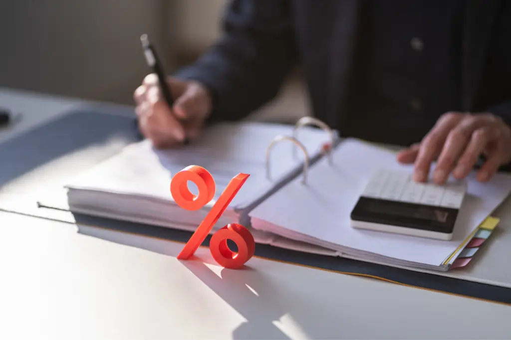 Finansal analiz ve reel faiz hesaplamaları bağlamında, bir masa üzerinde açık bir defterdeki belgelere not alan ve hesap makinesi kullanan kişinin elleri ile ön planda duran belirgin turuncu yüzde işareti.