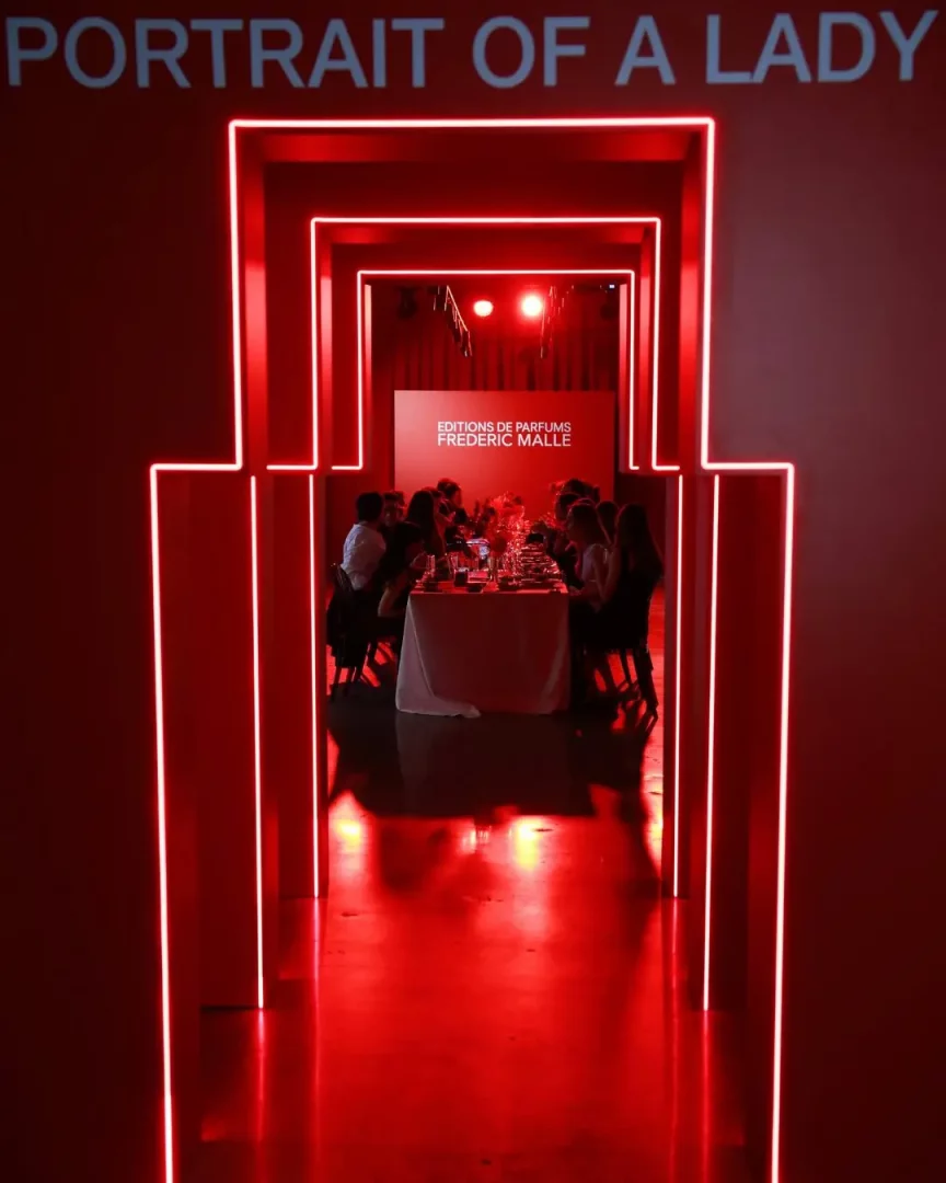 Kırmızı ışıkların ve neon çerçevelerin oluşturduğu etkileyici bir koridorun sonunda, Editions de Parfums Frederic Malle logosunun yansıtıldığı bir ekrana doğru oturan misafirlerin katıldığı, markanın sofistike atmosferini yansıtan özel bir Frederic Malle etkinliği.