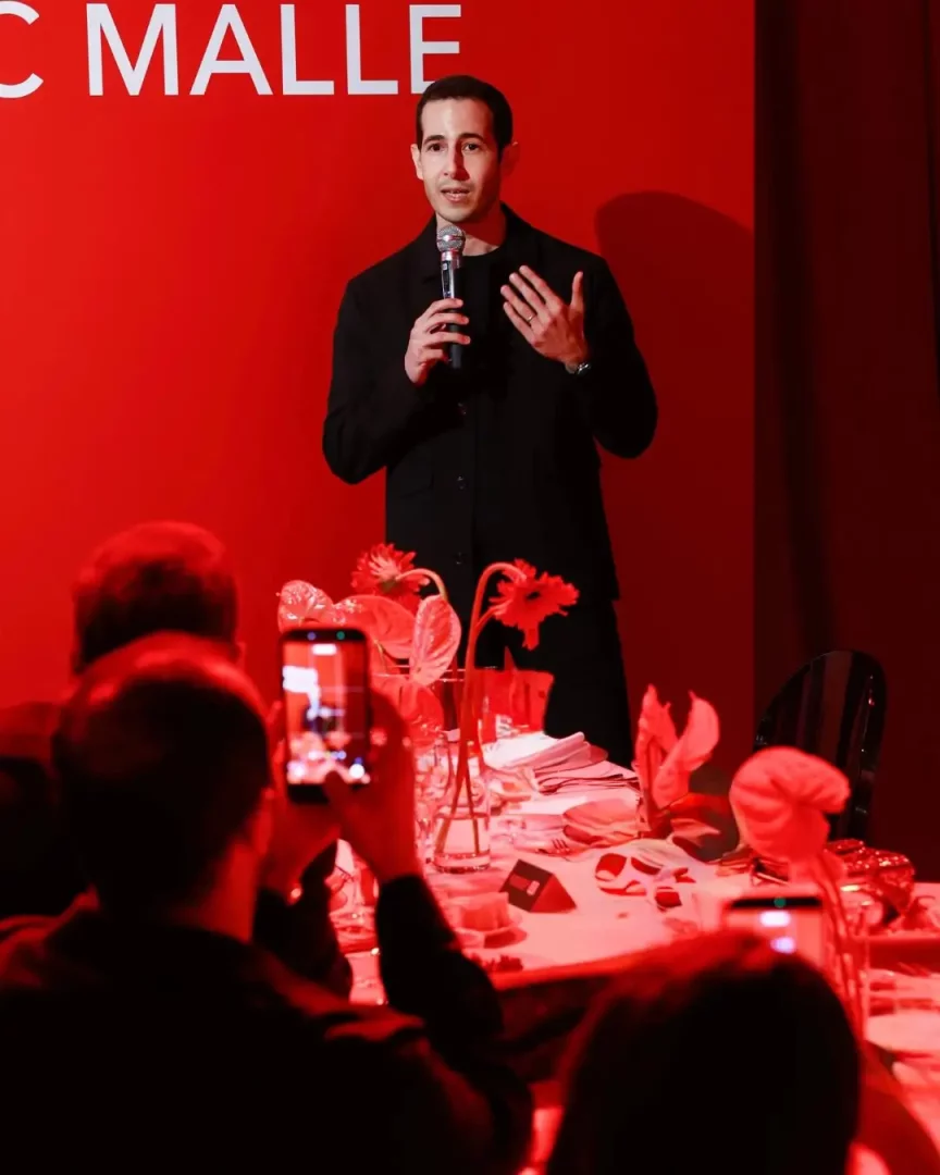 Frederic Malle etkinliğinde, kırmızı tonlarda aydınlatılmış bir sahnede, siyah giysili bir adamın mikrofonla konuşarak el hareketi yaptığı ve masalarda oturan konukların telefonlarıyla bu özel anı kayıt altına aldığı, zarif çiçek aranjmanlarıyla süslü bir ortam.