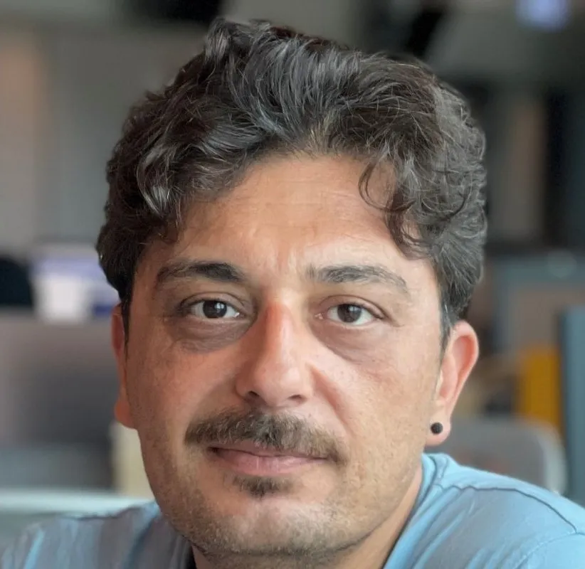 Olkan Özyurt, koyu kıvırcık saçları, bıyığı ve hafif sakallı yüzüyle kameraya doğrudan bakarken hafifçe gülümseyen, göz altlarında belirgin morluklar olan, sol kulağında küçük siyah bir küpe ve üzerinde açık mavi bir tişört bulunan yakından çekilmiş bir portrede görülüyor.