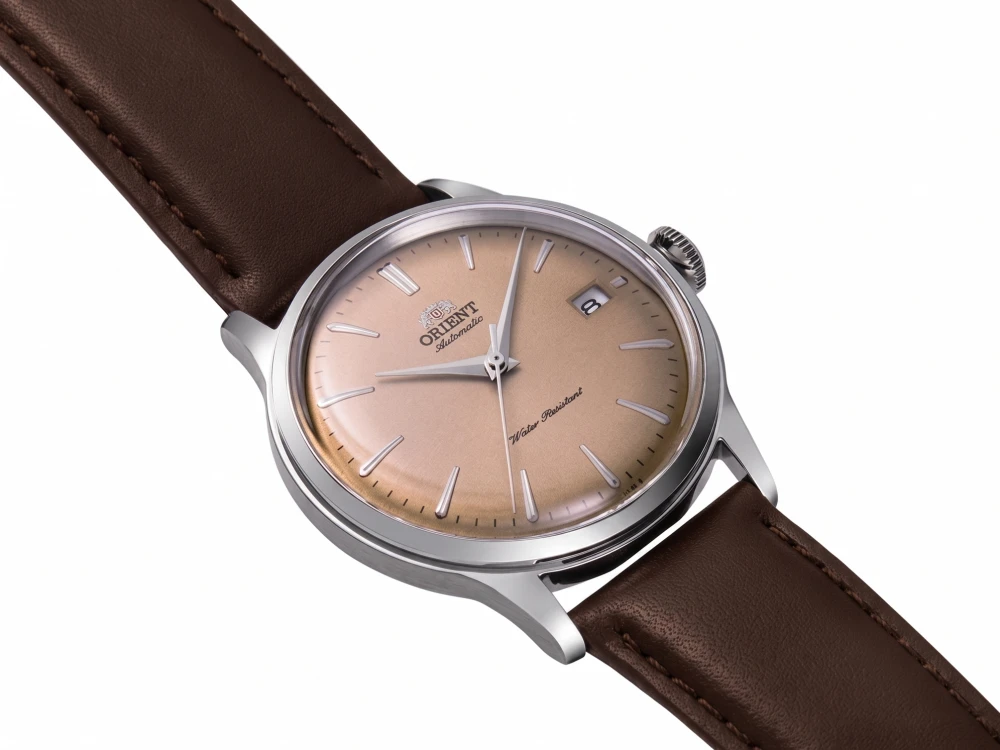 Açık kahverengi kadranlı, gümüş renkli kasalı ve kahverengi deri kayışlı bir Orient Bambino otomatik kol saati, kadranında Water Resistant yazısı ve 3 yönünde tarih penceresi net bir şekilde görülüyor.