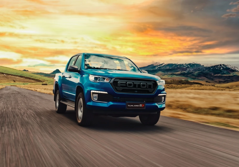 Otokar Foton Tunland G7 mavi pickup, dinamik bir açıyla uzanan asfalt yolda hızla ilerlerken, arkasında karlı zirvelere sahip dağlar ve turuncu-sarı renklerin hakim olduğu etkileyici bir gün batımı gökyüzüyle çevrili bir manzara sunuyor.