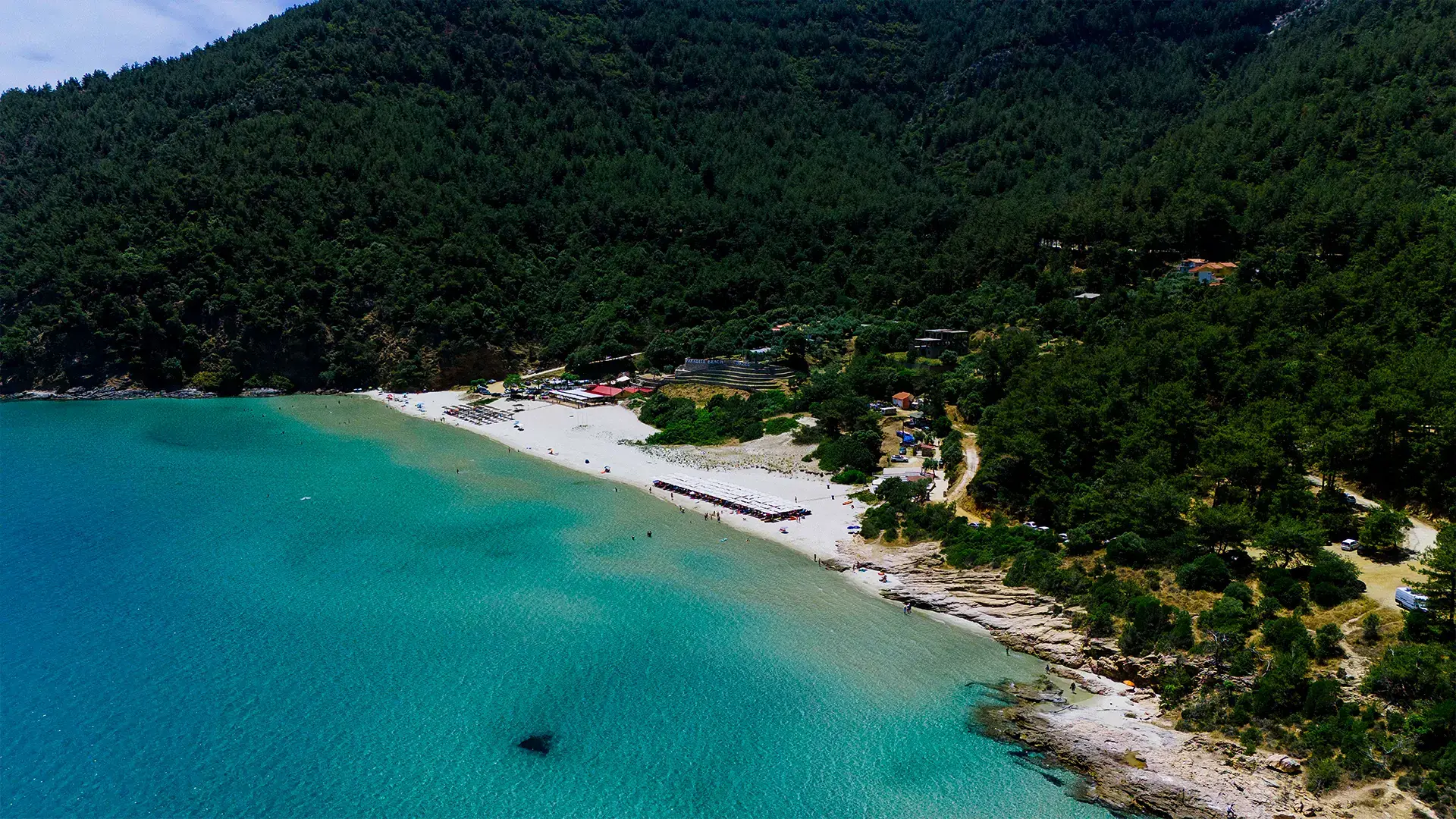 Cennet Plajı'nın havadan çekilmiş görüntüsünde, berrak turkuaz sularla çevrili beyaz kumlu sahil boyunca sıralanmış şezlonglar ve şemsiyeler altında dinlenen insanlar, arka planda yemyeşil sık ormanlık bir dağ yamacı ve plaj tesisleri dikkat çekiyor.