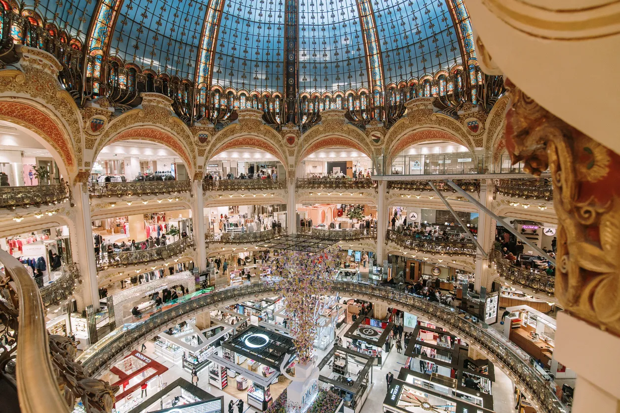 Paris'in simgesel alışveriş duraklarından Galeries Lafayette'in Art Nouveau tarzı vitraylı görkemli kubbesi altında, katlar boyunca uzanan süslü balkonlarda alışveriş yapan kalabalıklar ve lüks mağaza standları görülüyor.
