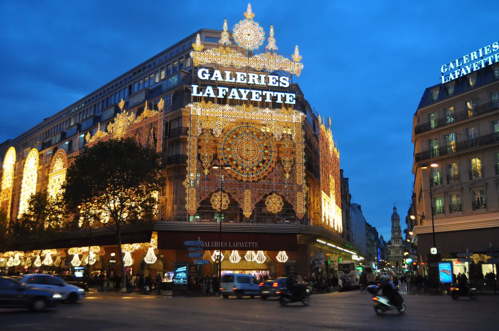 Paris'in simge yapılarından Galeries Lafayette, akşam karanlığında dış cephesini baştan aşağı süsleyen altın rengi ışıklı süslemeleri ve ortadaki görkemli gül pencere motifleriyle ışıl ışıl parlayarak büyüleyici bir şehir manzarası sunarken, önündeki işlek caddede hareketli bir kalabalık ve trafikteki araçlar dikkat çekiyor.