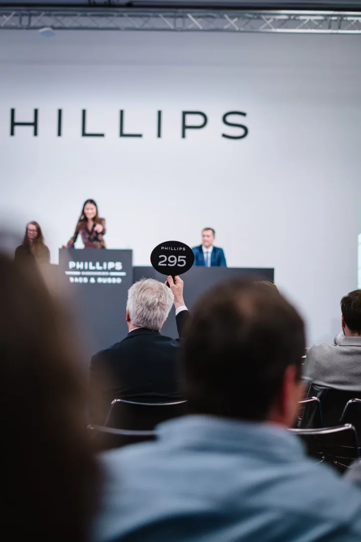 Gri saçlı bir katılımcı, arka planda Phillips logosunun ve müzayede görevlilerinin seçildiği bir salonda, elinde 295 numaralı teklif kartını havaya kaldırarak Phillips Cenevre saat müzayedesine aktif olarak katılıyor.