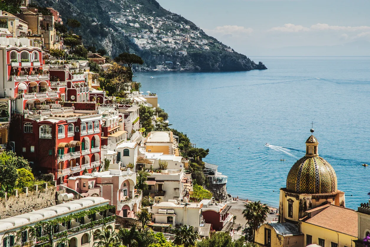 Positano, İtalya'nın masmavi denize dik inen yemyeşil yamaçlarına kurulu, kırmızı, beyaz ve sarı renkli binalarıyla dikkat çeken, denize nazır kendine özgü desenli çini kubbeli kilisesi ve uzaktan görünen tekneleriyle pitoresk bir sahil kasabasının güneşli bir gündeki genel görünümü.