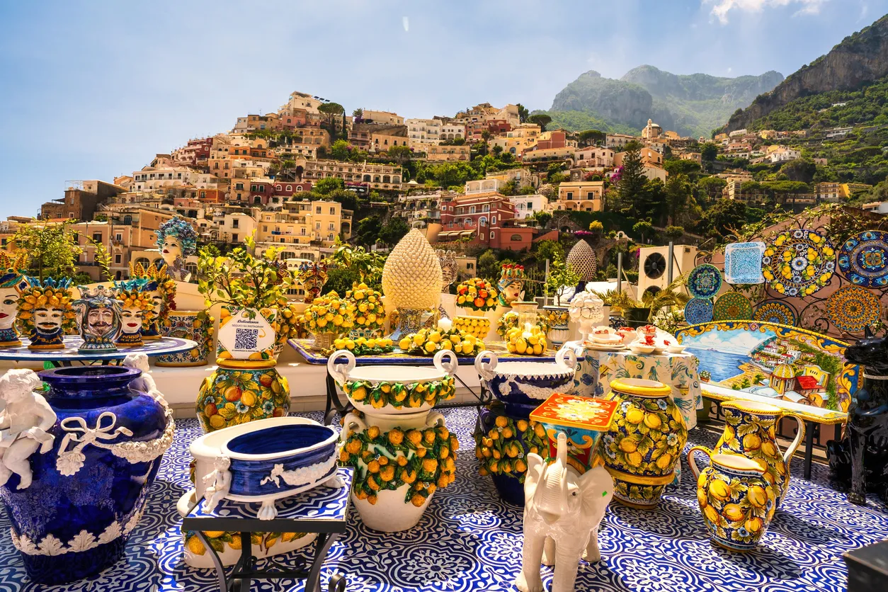 Positano'da, limon desenli seramik vazoların, tabakların ve maskelerin sergilendiği canlı bir açık hava tezgahının önünde, arka planda yamaçlara sıralanmış pastel renkli evler ve dağların nefes kesen manzarasıyla bu eşsiz destinasyonun ruhunu yansıtan bir görsel.