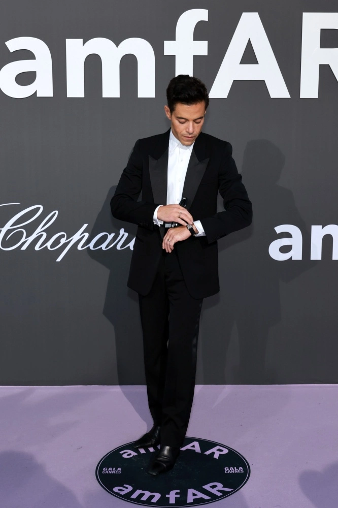 Rami Malek, siyah smokini ve beyaz gömleğiyle, amfAR ve Chopard logolarının olduğu gri bir fonun önünde mor halıda dururken sol bileğindeki Cartier Tank saatine bakıyor, Cannes kırmızı halı etkinliğindeki şıklığını sergiliyor.