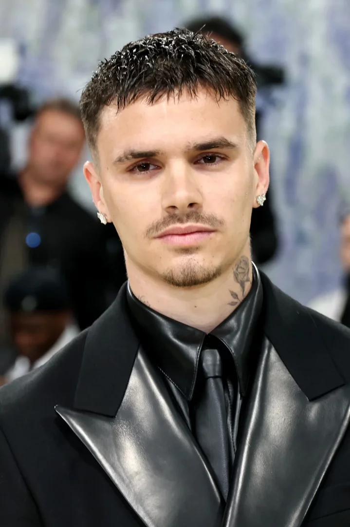 Romeo Beckham, Met Gala etkinliğinde çekilmiş bu yakın planda, siyah parlak deri yakalı bir takım elbise, deri gömlek ve kravat giymiş, kısa saçları, hafif sakalları, kulağında parlayan küpeleri ve boynundaki gül dövmesiyle dikkat çekiyor.