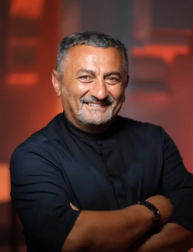 Sabit Akkaya, koyu renk, yakasız bir gömlek giymiş, kollarını kavuşturmuş ve kameraya doğru sıcak bir gülümsemeyle poz veriyor; kısa gri saçları ve gri sakalları, hafif kırmızı tonlardaki bulanık arka plan önünde belirginleşiyor.