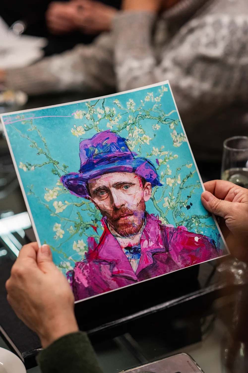 Canlı renklerle dolu, mor bir şapka ve fuşya bir ceket giymiş, turkuaz renkli ve çiçek açmış dallarla süslü bir arka plana sahip, Van Gogh'a benzeyen sakallı bir adamın dışavurumcu portresi, iki el tarafından tutulurken yakından görüntüleniyor.