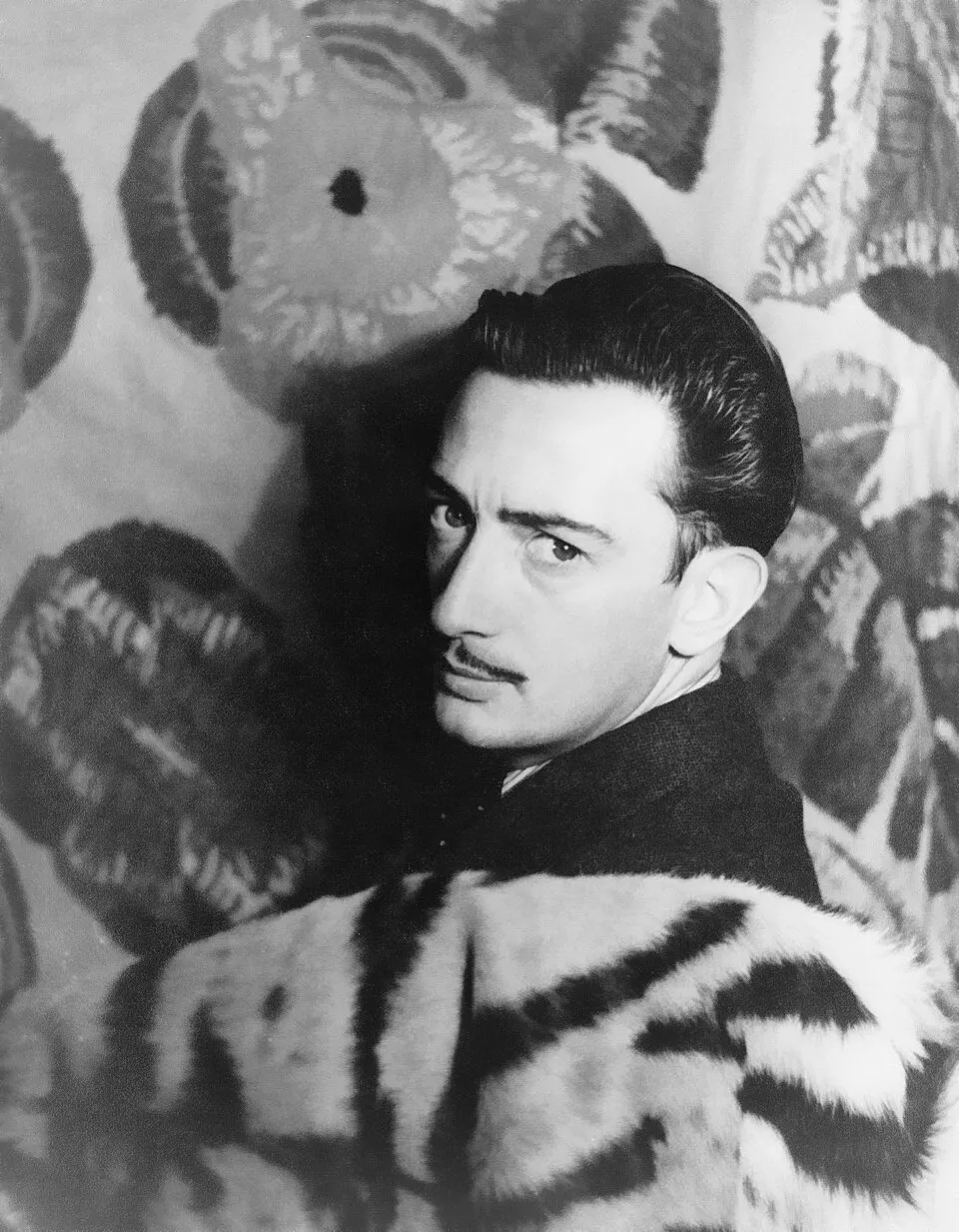 Siyah beyaz bir portre fotoğrafında, bıyıklı ve geri taranmış saçlı, tanınmış sürrealist sanatçı Salvador Dalí, çizgili kürk detaylı bir kıyafetle, arka plandaki çiçekli desenli kumaşın önünde yoğun bir bakışla doğrudan izleyiciye dönük poz veriyor.