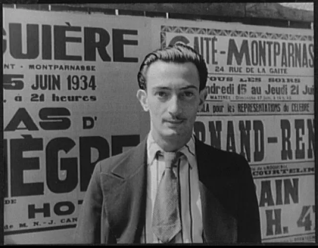 Siyah beyaz bir fotoğrafta, genç Salvador Dalí, 1934 tarihli Fransızca reklam posterlerinin önünde, karakteristik ince bıyığı ve arkaya taranmış saçlarıyla kameraya doğrudan bakarak gençlik yıllarından etkileyici bir portre sunuyor.