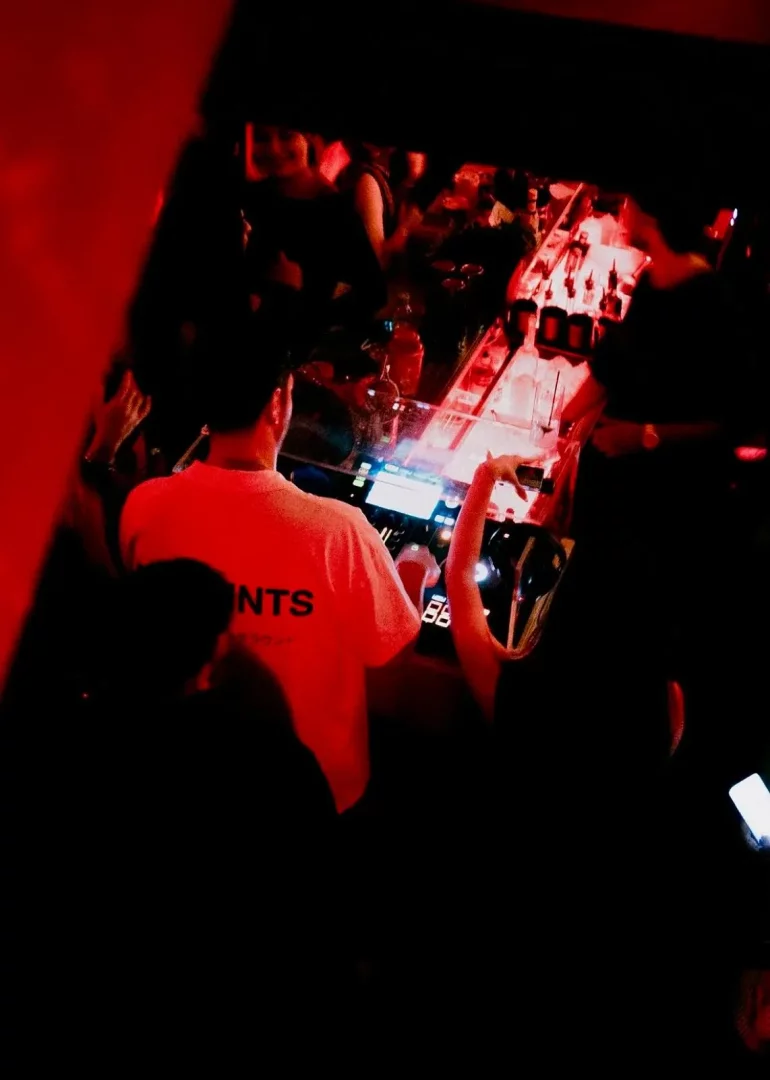 Scatola Kuruçeşme'de kırmızı ışıklar altında bir DJ'in elektronik müzik ekipmanlarıyla performans sergilediği, çevresinde dans eden veya sohbet eden insanların bulanık silüetlerinin seçilebildiği dinamik bir gece kulübü sahnesi.