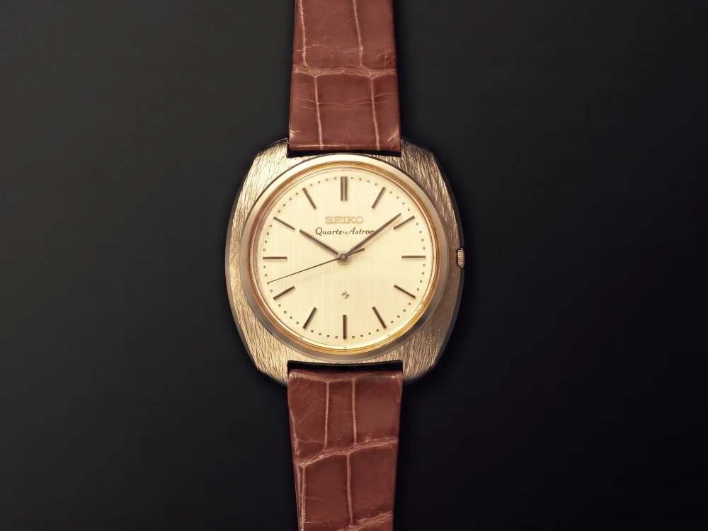 Dikdörtgenimsi altın kasalı, krem kadranlı ve kahverengi timsah derisi kayışlı, saatçilik tarihinde bir dönüm noktası olan ikonik Seiko Quartz Astron, ilk quartz saat olarak koyu bir zemin üzerinde dikey bir şekilde sergilenmektedir.