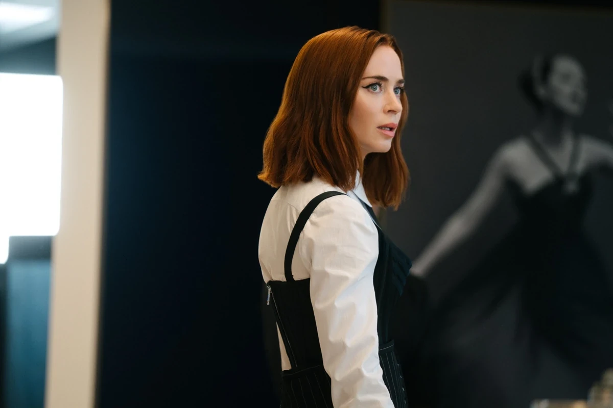Şeytan Marka Giyer filminden Emily Blunt, omuz hizasında kızıl kahve dalgalı saçları, dikkat çekici makyajı ve beyaz gömlek üzerine giydiği siyah, çizgili, korse tarzı askılı üstüyle, bir ofis moda sahnesinde şaşkın bir ifadeyle yana doğru bakıyor, arkasında flu bir kadın silueti beliriyor.
