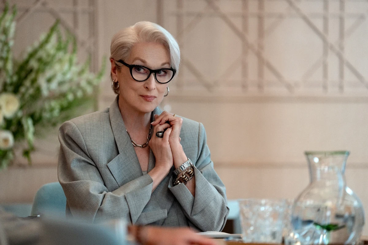 Meryl Streep'in Şeytan Marka Giyer filmindeki ikonik Miranda Priestly karakterini canlandırdığı bu karede, kısa beyaz saçlı, siyah kedi gözü gözlüklü, gri blazer ceketli aktris ellerini kenetlemiş bir toplantı masasında ciddi ve dikkatli bir ifadeyle izleyiciye bakıyor.