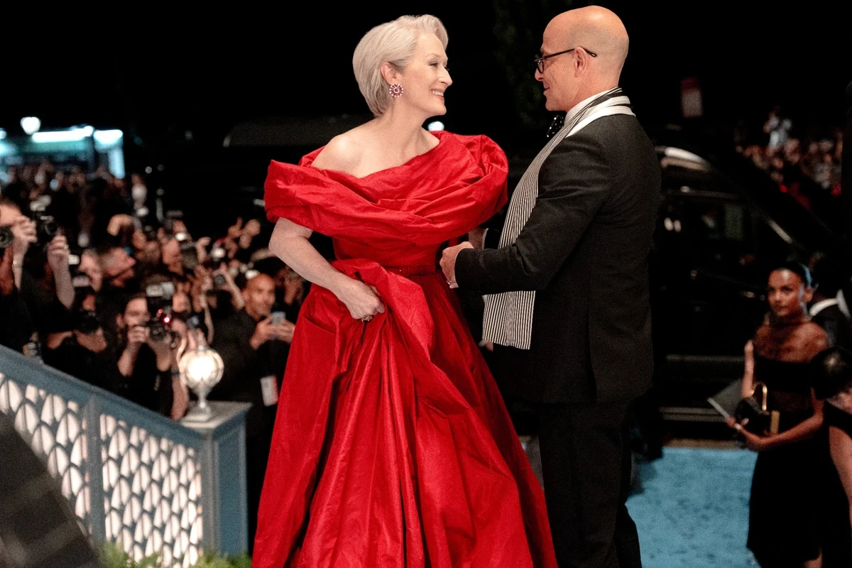 The Devil Wears Prada 2 filmindeki ikonik kırmızı halı sahnesinden alınan bu karede, Miranda Priestly göz kamaştırıcı kırmızı, omuzları açık bir elbiseyle neşeyle gülümserken, yanında zarif siyah takımı ve çizgili fularıyla Nigel Kipling ona bakıyor ve arka planda gazeteciler onları fotoğraflıyor.
