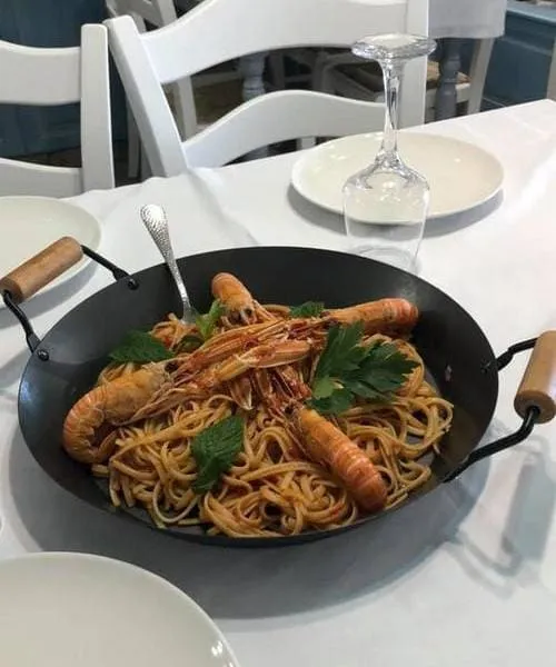 Beyaz masa örtülü bir Simi restoranında servis edilen, siyah tavada bol porsiyonlu, taze otlarla süslenmiş, domates soslu makarnanın üzerinde büyük karideslerin yer aldığı iştah açıcı bir deniz mahsulleri yemeği sunumu.