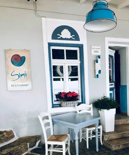 Simi restoranının dış cephesinde, beyaz badanalı duvarda mavi çerçeveli şirin bir pencerenin önünde, açık mavi masa ve hasır oturaklı iki beyaz sandalyenin bulunduğu bir oturma alanı görülüyor; üstte asılı turkuaz renkli lamba ve pencere kenarındaki kırmızı çiçekler Akdeniz'in sıcak atmosferini yansıtıyor.