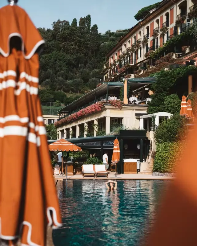 Ön plandaki turuncu şemsiyelerin arasından, yemyeşil tepelere yaslanmış, balkonları pembe çiçeklerle süslü zarif Splendido Oteli'nin teraslı restoranlarını ve havuzunda rahatça yüzen bir misafiri içeren, Portofino'nun lüks ve dinlendirici ambiyansını yakalayan bir manzara.