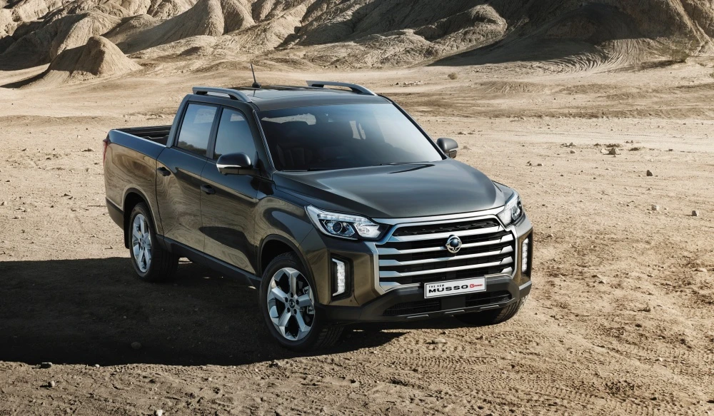 Arka planda engebeli, kahverengi dağların ve kumlu zeminin görüldüğü, kurak bir arazide duran, koyu gri renkli yeni SsangYong Musso Grand pickup kamyonet, macera dolu off-road sürüşleri için hazır bir şekilde konumlanmış.