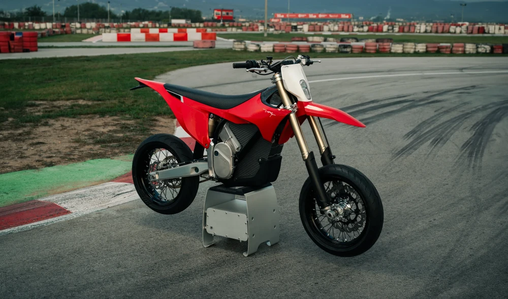 Canlı kırmızı renkteki Stark Varg MX-12 elektrikli arazi motosikleti, asfalt üzerinde belirgin lastik izlerinin görüldüğü bir yarış pistinde, gümüş renkli bir sehpa üzerinde dururken, arka planda kırmızı beyaz bariyerler ve hafif sisli dağlar uzanıyor.