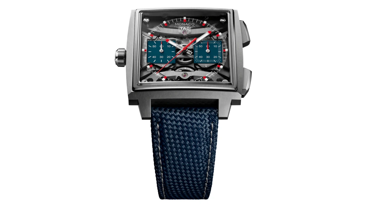 Ortasında beyaz bir arka plan üzerinde duran, iskelet kadranı ve mavi kronograf alt kadranlarıyla mekanizmasını sergileyen, modern kare kasalı TAG Heuer Monaco Evergraph kronograf saat, lacivert dokulu kayışı ve kırmızı ibreleriyle dikkat çekiyor.