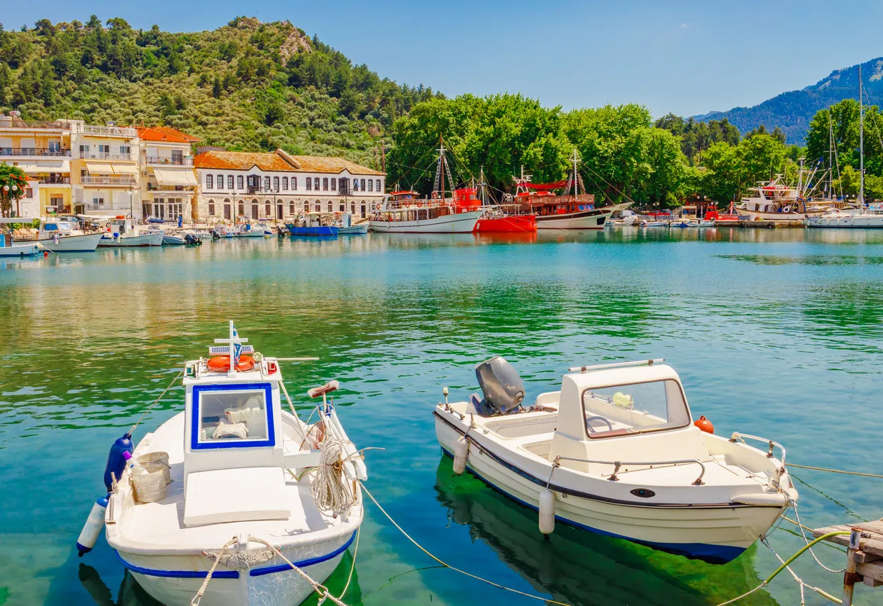 Thassos'un canlı limanında, masmavi berrak suda demirlemiş çok sayıda teknenin önünde, beyaz ve mavi şeritli küçük bir tekne ile yanındaki beyaz motorlu tekne ön planda dikkat çekerken, kıyıdaki geleneksel mimariye sahip binalar ve yemyeşil tepeler adanın çekici atmosferini yansıtmaktadır.
