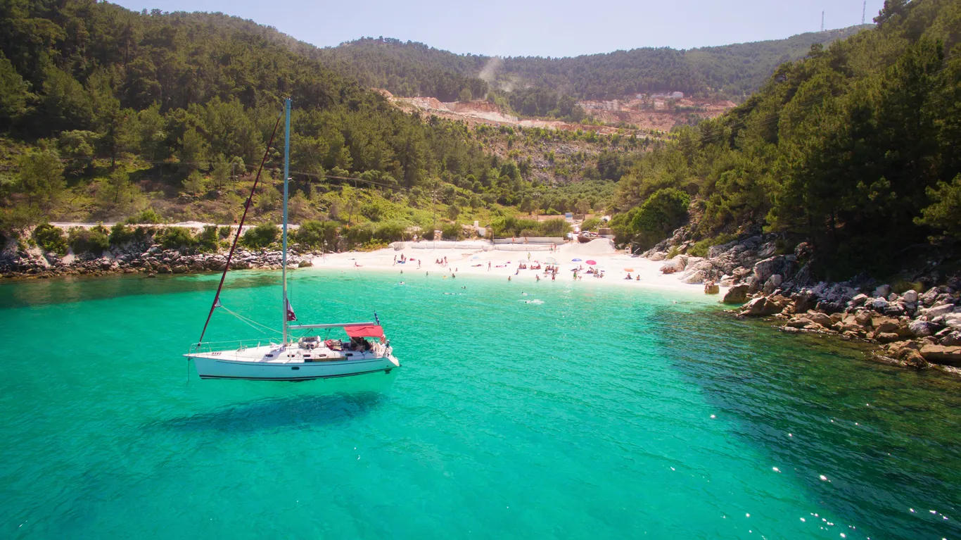 Thassos adasının eşsiz doğal güzelliklerini sergileyen bu fotoğrafta, turkuaz renkli berrak sularında hafifçe salınan beyaz bir yelkenli tekne ve arka planda yemyeşil çam ormanlarıyla çevrili kalabalık, güneşli bir kumlu plaj canlanıyor.