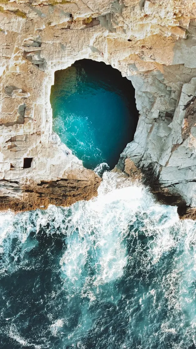 Thassos'un derin mavi sularının beyaz köpüklerle kayalara vurduğu, antik çağlardan kalma bir gizli tarih barındıran dramatik uçurumlar arasındaki turkuaz renkli deniz mağarasının büyüleyici kuşbakışı görünümü.