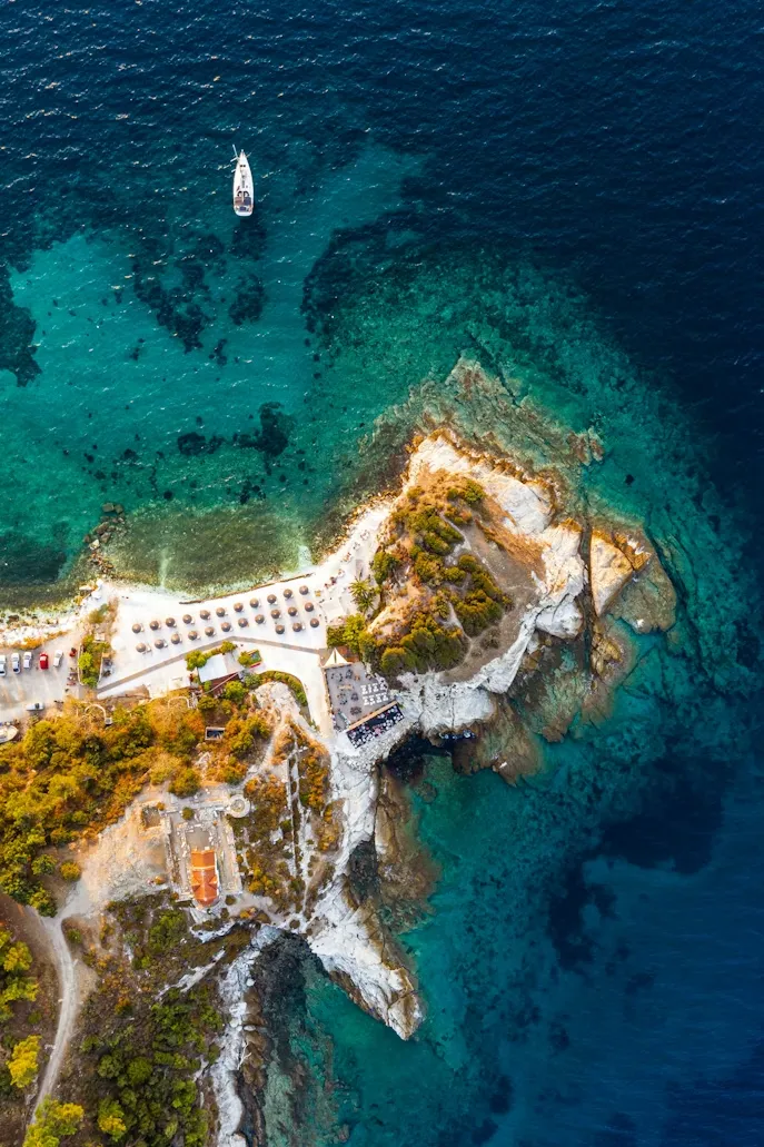 Thassos kıyılarının kuşbakışı görünümünde, berrak turkuaz sularında süzülen beyaz bir yelkenli, şemsiyeli kumlu bir plaj, kayalık burun üzerinde yer alan bir tesis ve antik dönemden kalma gibi görünen kiremit çatılı bir yapının bulunduğu sahil şeridi, adanın gizli tarihini yansıtan büyüleyici bir manzara sunuyor.