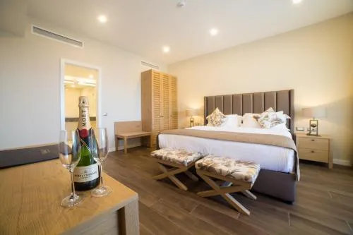 Thassos'taki lüks bir konaklama deneyimini yansıtan bu modern otel odasında, minderli başlığı olan büyük bir yatak, önünde çiçek desenli iki puf, açık renk ahşap mobilyalar ve zarif aydınlatmalar bulunuyor; ön planda ahşap bir masa üzerinde ise bir şişe Moët & Chandon şampanya ve iki kadeh, davetkar bir atmosfer sunuyor.