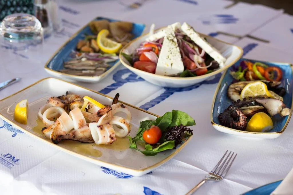 Thassos'un eşsiz mutfağını yansıtan bu canlı sofrada, özenle hazırlanmış ızgara kalamar halkaları, bol beyaz peynirli taze Yunan salatası, iştah açıcı ızgara ahtapot ve çeşitli deniz mahsulleri mezeleri ile dolu, Akdeniz esintili bir ziyafet sunuluyor.