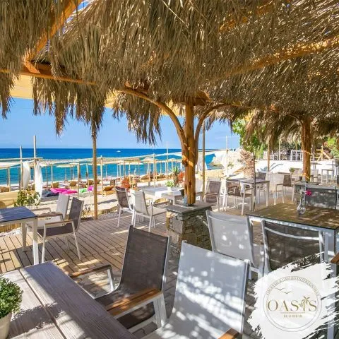 Thassos'taki Oasis plaj barının hasır çatılı, ahşap zeminli açık hava oturma alanında, modern sandalyelerin ve masaların denize nazır konumlandığı, arka planda uzanan kumlu plajın şezlonglar ve hasır şemsiyelerle dolu olduğu, masmavi denizin huzur veren görüntüsüyle buluşan davetkar bir atmosfer.