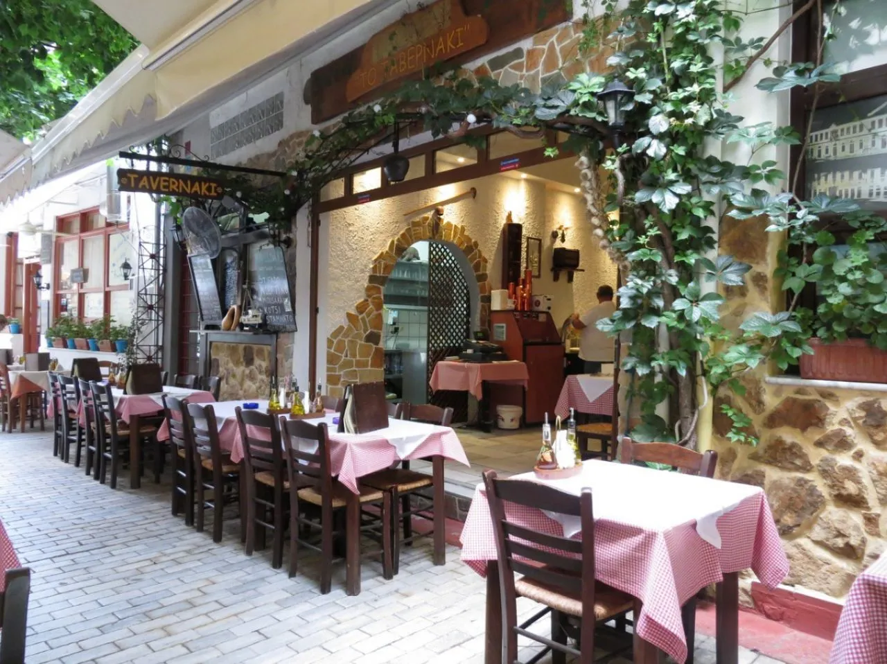 Yemyeşil sarmaşıklarla kaplı taş ve beyaz duvarlara sahip Thassos'taki Tavernaki adlı restoranın dış yemek alanında, kırmızı-beyaz kareli masa örtüleri ve ahşap sandalyelerle donatılmış çok sayıda masa, keyifli bir yemek deneyimi için hazır beklemektedir.