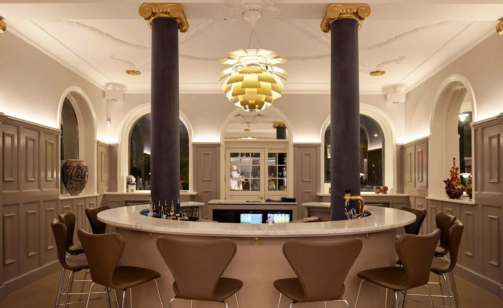 Kopenhag'daki The Samuel restoranının sofistike bar alanında, altın rengi, katmanlı bir avizenin aydınlattığı beyaz mermer bar tezgahının etrafında dizili kahverengi deri bar tabureleri ve altın başlıklı sütunlar, modern ve klasik unsurları bir araya getiren şık bir atmosfer yaratıyor.