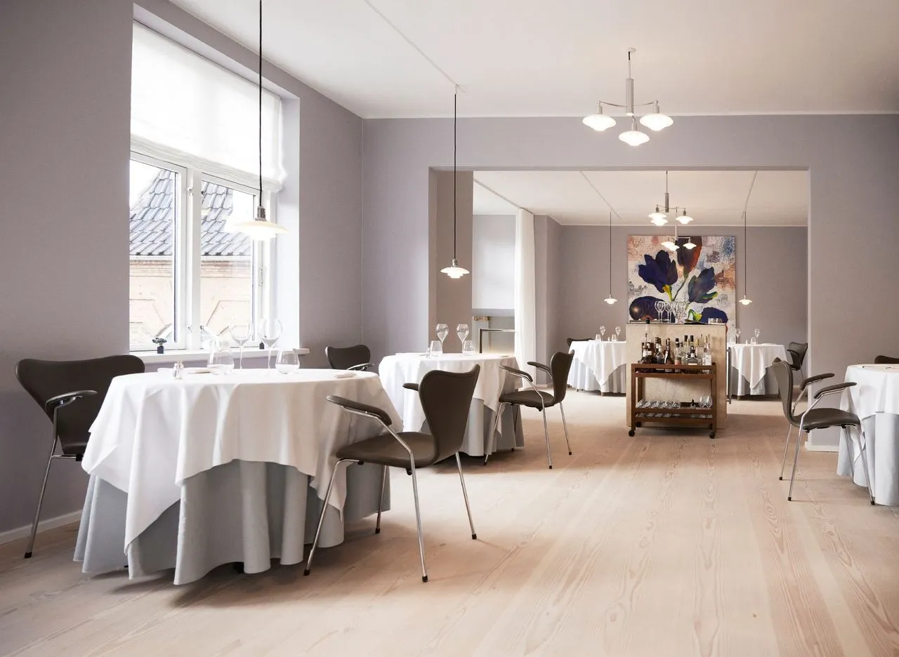 Aydınlık ahşap zemine ve açık gri duvarlara sahip, Kopenhag'da bulunan modern bir restoranın iç mekanı, beyaz örtülü şık masaları, koyu renk sandalyeleri ve arka planda büyük bir soyut çiçek tablosu ile zarif bir yemek deneyimi sunuyor.