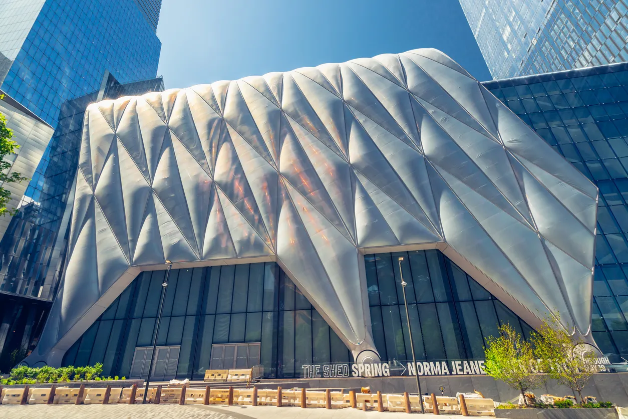 New York'un mimari yapısına özgü modern bir estetiği sergileyen, parlak elmas desenli gümüş renkli dış cephesi ve altında The Shed Spring → Norma Jeane yazısı bulunan The Shed binası, mavi gökyüzü altında parıldayan cam kulelerin arasında yükseliyor.