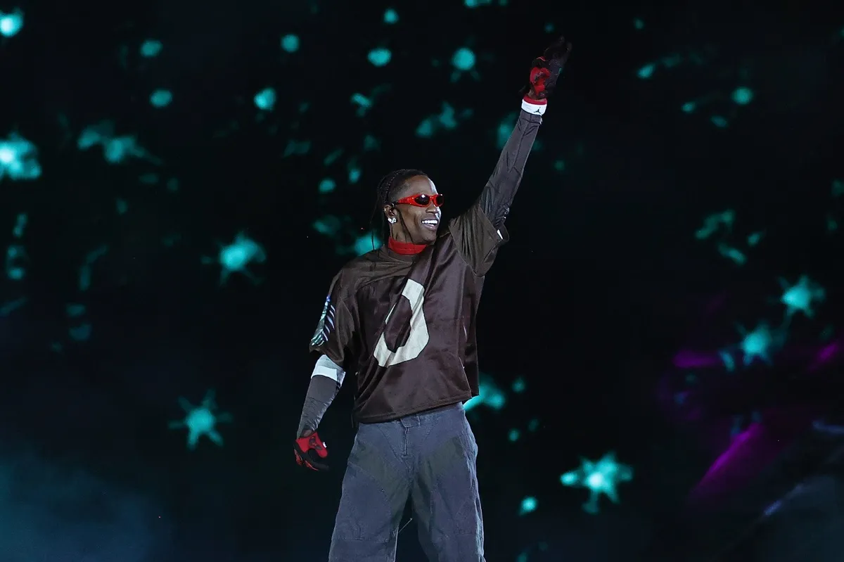 Travis Scott, hip-hop sahnesindeki yoğun enerjisini kırmızı güneş gözlükleri ve eldivenleriyle yansıtırken, arka plandaki parlayan yıldız benzeri ışıklar eşliğinde sağ kolunu havaya kaldırarak dinleyicileriyle coşkulu bir an paylaşıyor.