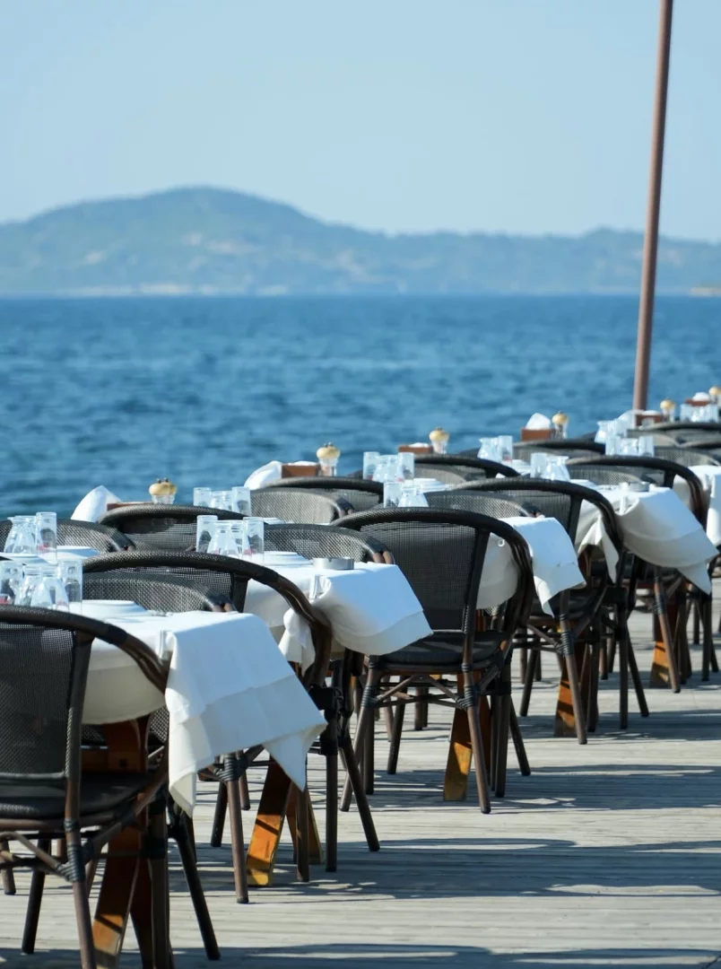 Urla sahilindeki bir işletmede, masmavi denize sıfır ahşap bir iskele üzerinde güneşin altında sıra sıra dizilmiş, beyaz örtülü, boş masa ve hasır sandalyeler ile uzakta belli belirsiz dağ silüetleri yer alıyor.
