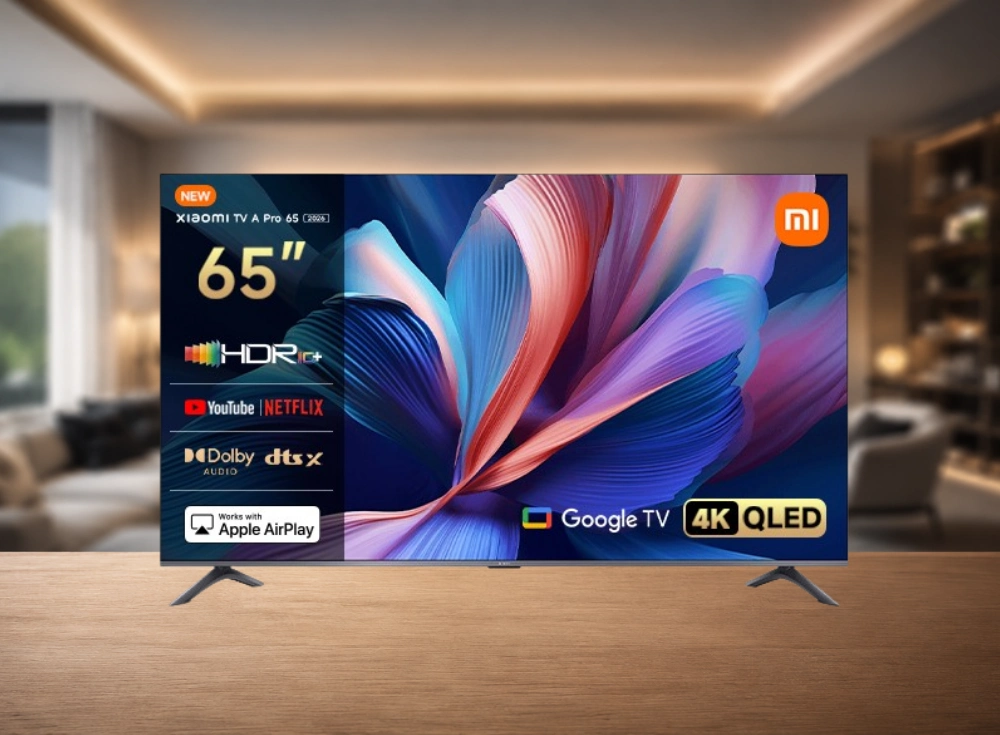 Ahşap bir zemin üzerinde duran, ince metal çerçeveli, canlı renklerde soyut bir duvar kağıdının gösterildiği, üzerinde Xiaomi TV A Pro 65 (2026), 65 inç, Google TV, 4K QLED, HDR10+, YouTube, Netflix, Dolby DTS X Audio ve Apple AirPlay logoları bulunan modern bir akıllı TV.