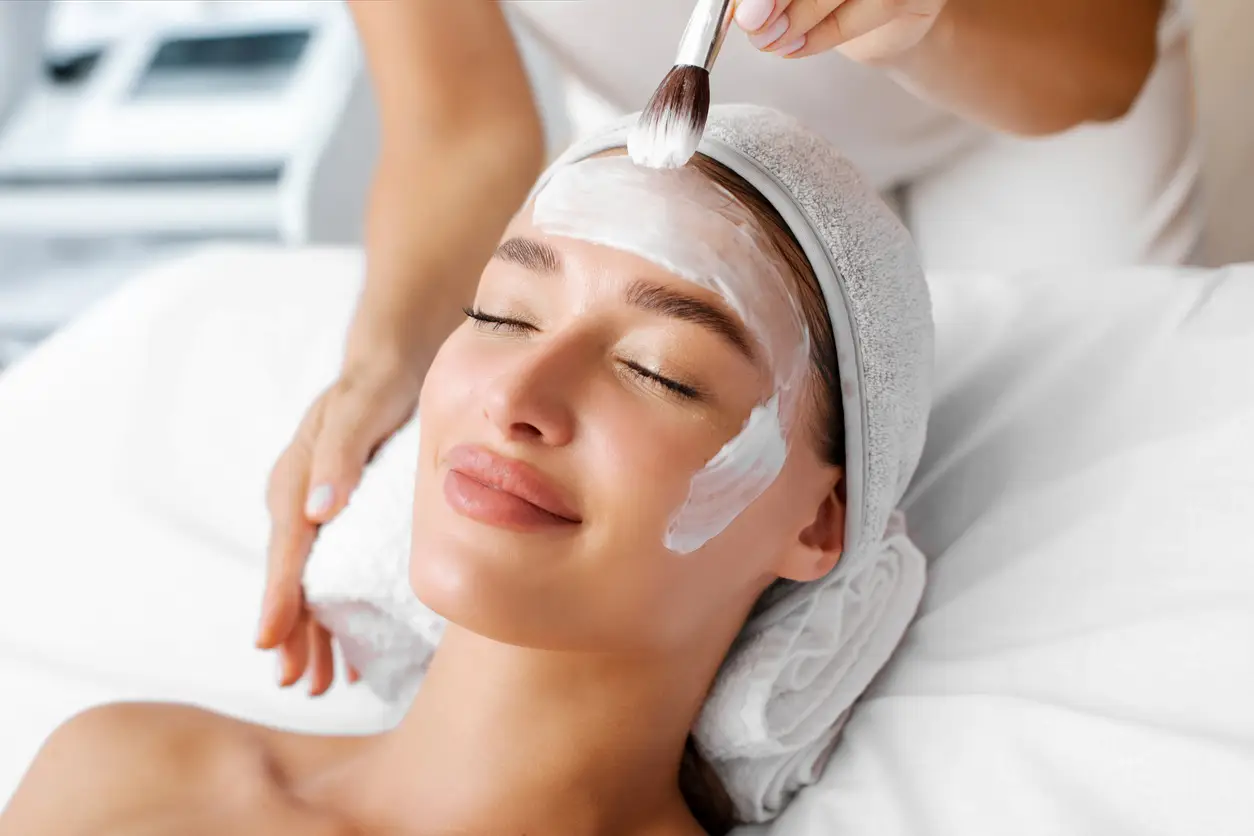 Beyaz bir havluyla sarılmış başı ve kapalı gözleriyle rahatlamış görünen bir kadının yüzüne, bir uzmanın fırça yardımıyla beyaz renkli bir maske uyguladığı bu görüntü, yazın tercih edilebilecek yenileyici klinik uygulamalardan birini anlatıyor.