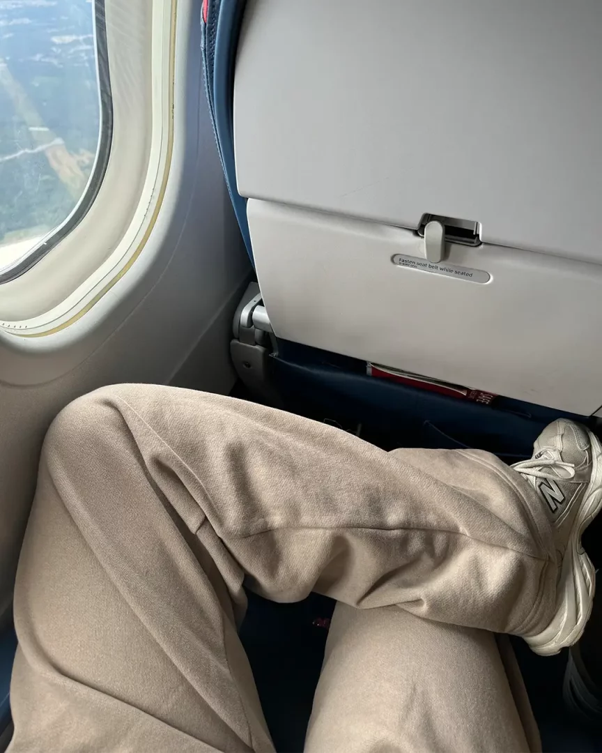 Bir uçak yolculuğu sırasında rahatlık arayan bir kişinin, açık bej rengi yumuşak kumaş pantolon ve spor ayakkabılar giydiği bacakları, pencere kenarında dinlenir bir şekilde duruyor; ön koltuğun arkasındaki katlanmış tepsi masası da görünüyor.
