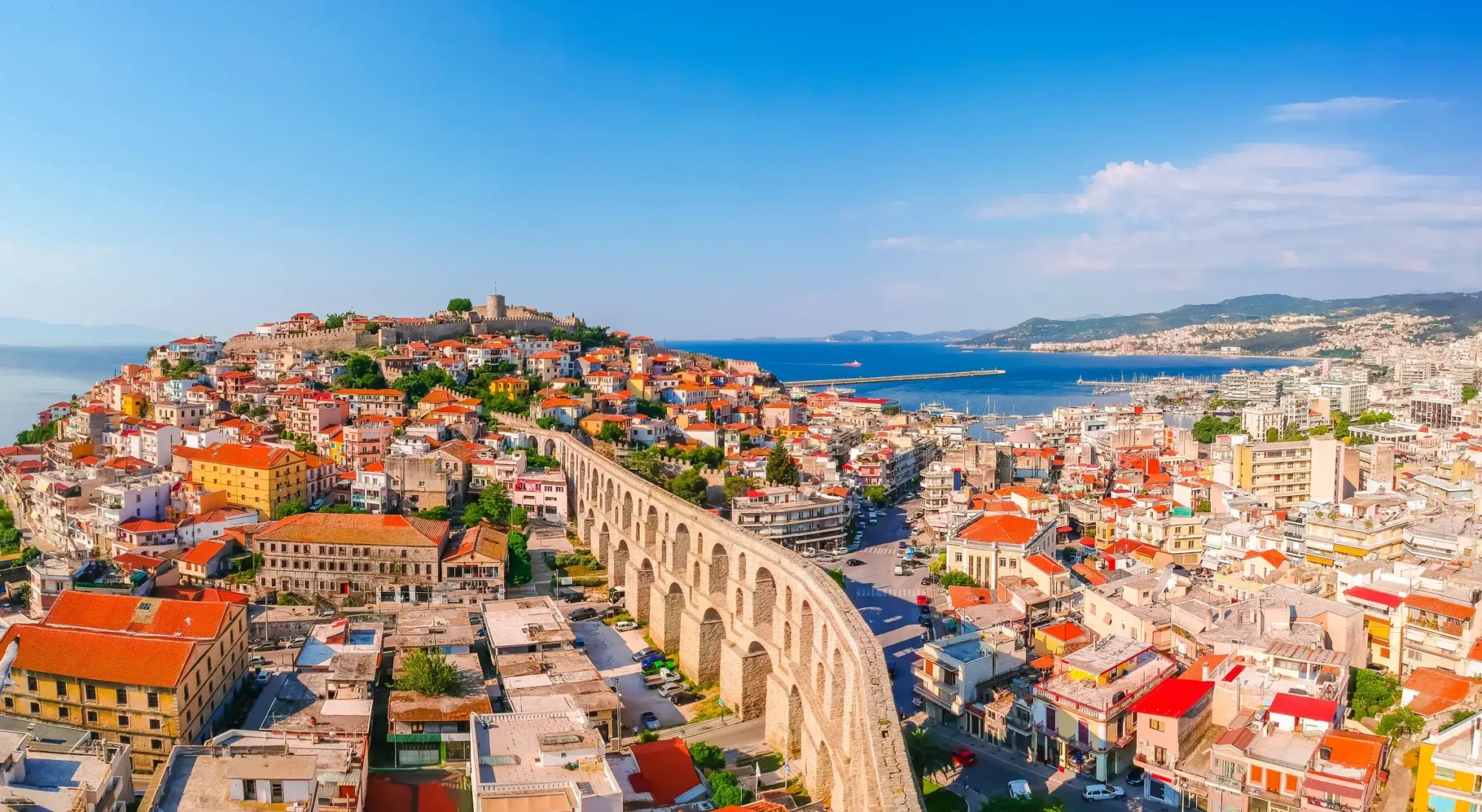 Yunanistan'ın masmavi Ege Denizi kıyısında, güneşli bir günde tepesinde tarihi bir kalenin yer aldığı, kemerli antik su kemerinin kırmızı ve renkli çatılı evlerin arasından geçerek uzandığı, limanda demirli teknelerin görüldüğü hareketli bir sahil şehri panoraması.