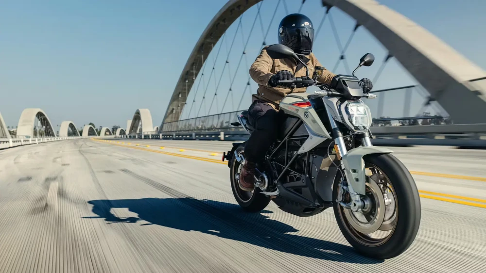 Güneşli bir günde, kemerli köprü üzerinde ilerleyen, kasklı ve kahverengi ceketli bir sürücünün kullandığı açık gri renkli Zero SR/F elektrikli naked motosiklet, yol boyunca uzun gölgelerle dinamik bir sahne oluşturuyor.