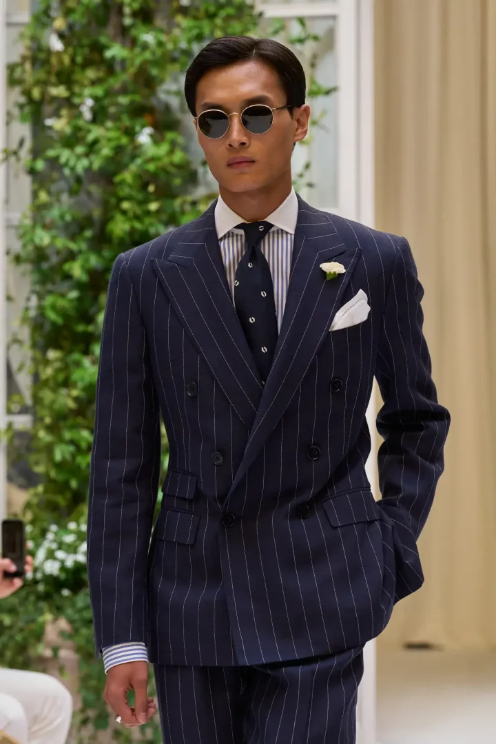 Lacivert ince çizgili kruvaze takım elbise, beyaz yaka çiçeği ve desenli kravatıyla şık görünen, yuvarlak güneş gözlüklü genç bir erkek model, Ralph Lauren İlkbahar 2027 erkek giyim koleksiyonundan modern ve klasik bir görünümü sergiliyor.